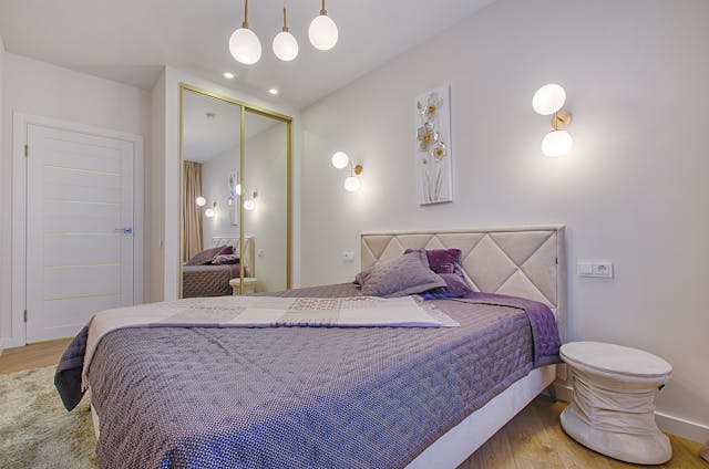 Spavaća soba sa ljubičastim prekrivačem na krevetu, dve zidne lampe i lusterom sa tri sijalice